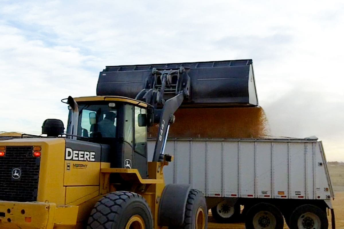Dymax Extra Wide Corn and Grain Bucket on a John Deere Wheel Loader John Deere wheel loader with a Dymax extra-wide corn and grain bucket dumping grain into a semi-trailer during harvest
