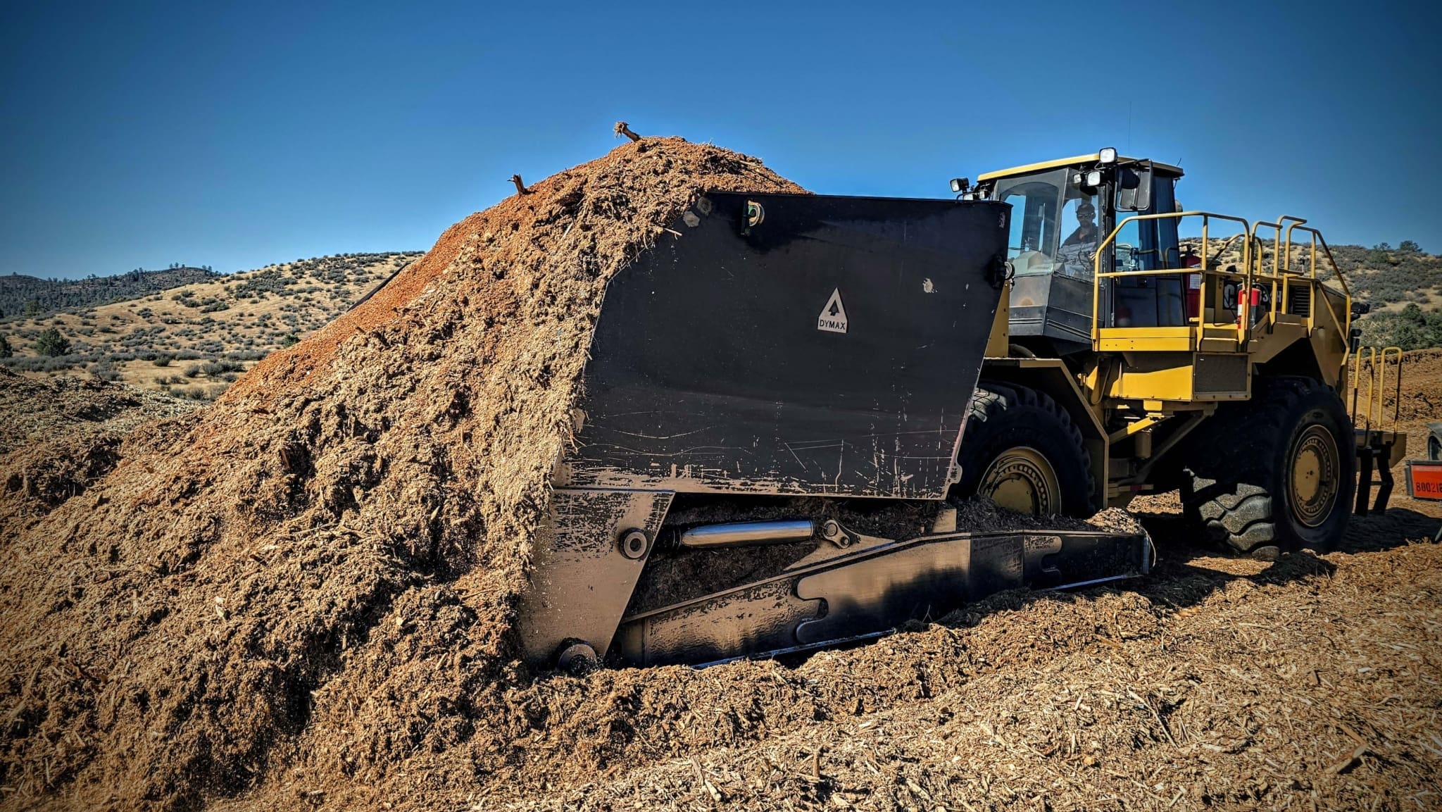 Dymax Wood Chip Scoop mounted on a Cat wheel dozer, filled to capacity while dozing a large wood chip pile at a biomass site
